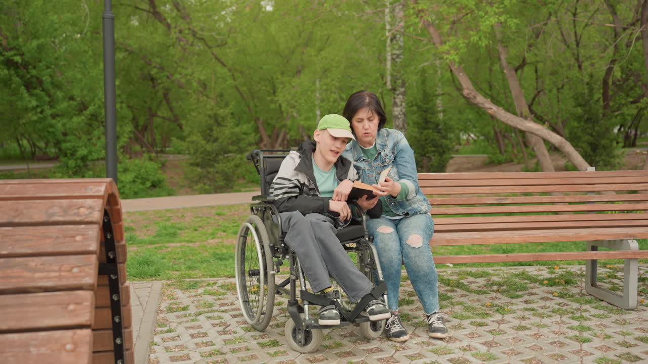 Woman And Young Man In Wheelchair Reading Book Together On Park Bench, Warm Supportive Bond, Woman Pointing At Pages While Both Smile, Spring Greenery Backdrop, Calm Companionable Leisure