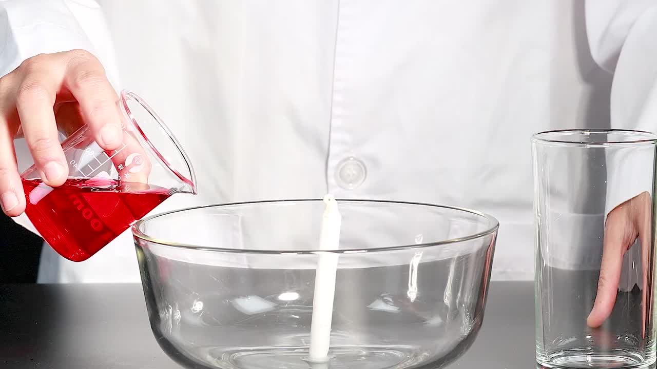 A person demonstrates a candle experiment with red liquid and glassware on a black surface.
