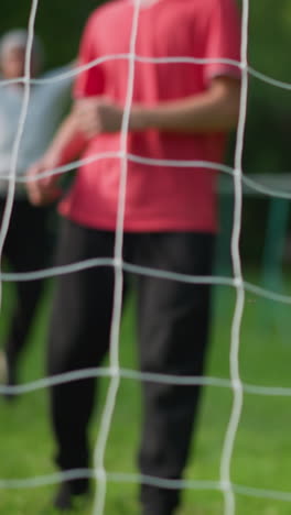 un primer plano de un niño con una camiseta roja dispara la pelota a la red, el portero pierde el salvamento, y un hombre en el fondo, borroso, se ve celebrando el gol