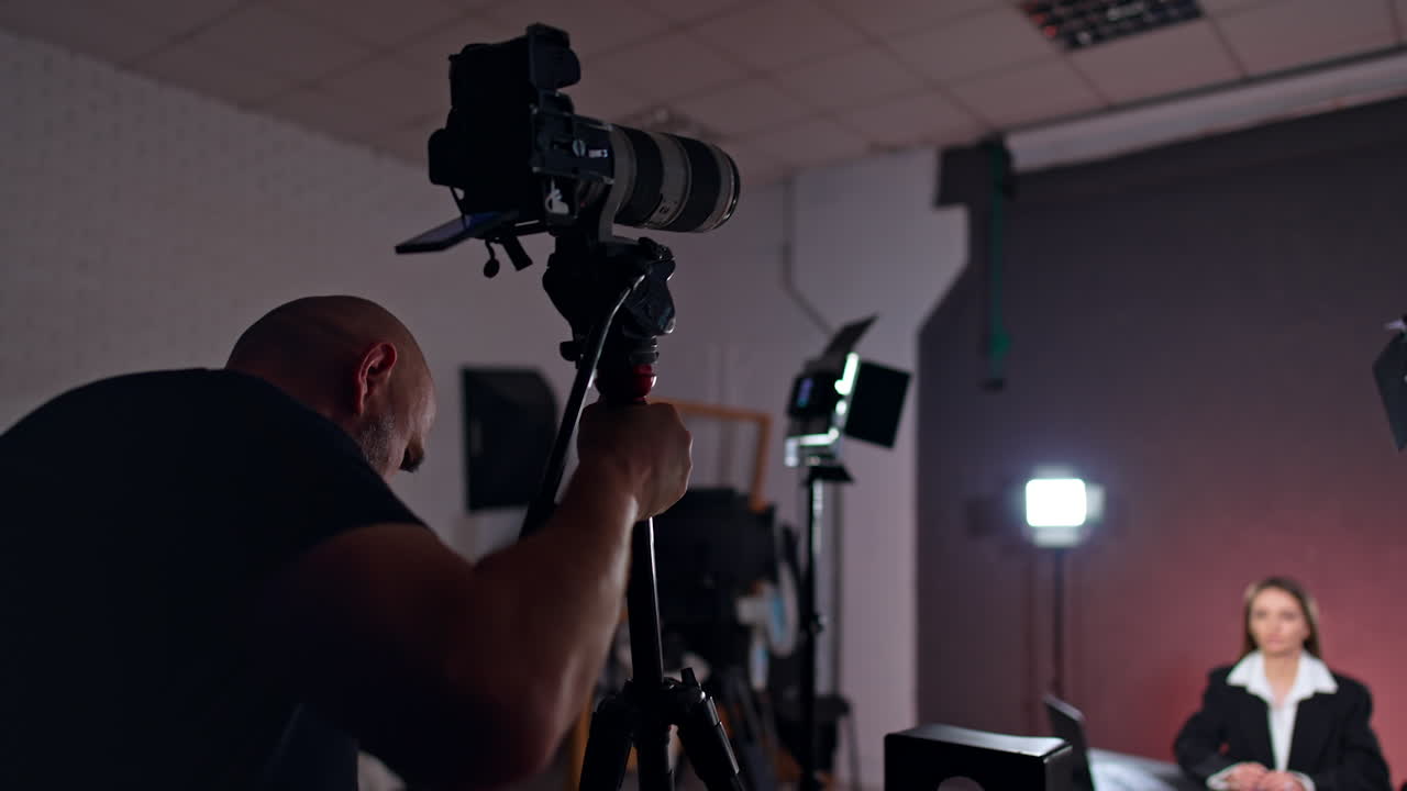 Professional cameraman setting camera in the studio. Female blogger sits at desk prepared for footage of blog.