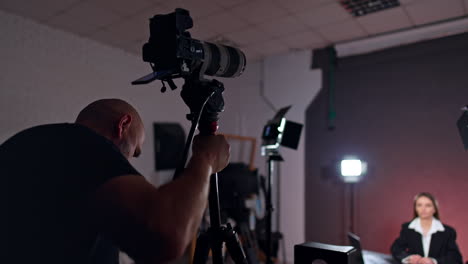 professional cameraman setting camera in the studio. female blogger sits at desk prepared for footage of blog.