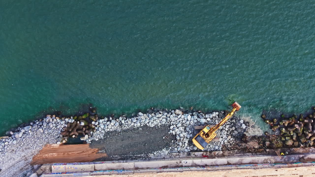 Lavori di costruzione sono in corso sul litorale mentre una macchina sposta rocce e sabbia vicino all'acqua cristallina alla luce del pomeriggio.