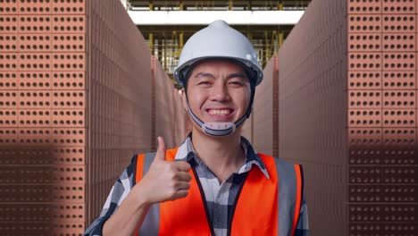 close up of asian male engineer with safety helmet smiling and showing thumbs up gesture to the camera while standing with red brick packed in stacks are stored