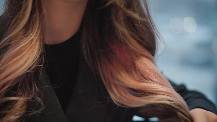 Persona che passa delicatamente le dita tra lunghi capelli castani con mèches mentre indossa un blazer scuro testurizzato e un top, creando una sottile arricciatura, volume e disposizione in una morbida luce interna con sfondo sfocato.