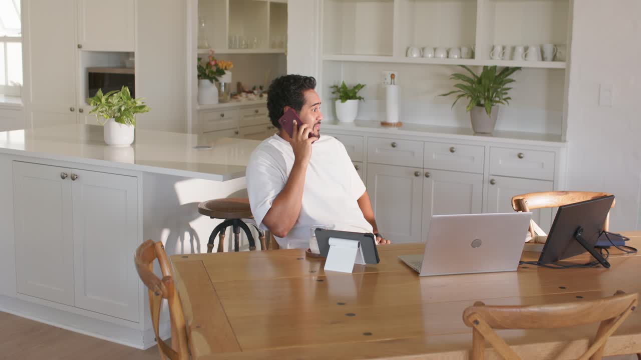 African American man answering work phone call, checking laptop and tablet at kitchen table