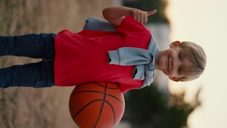 vertical video portrait of a happy little blond boy with blue eyes showing a like sign and sticking his thumb up while standing with a basketball in his hands. happy little boy has fun spending his leisure time actively playing sports games