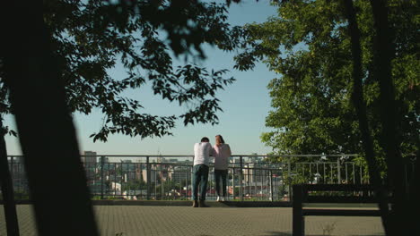 back view of couple standing and leaning on iron railing overlooking urban cityscape with the lady stand on the pavement surrounded by greenery and lit by warm sunlight