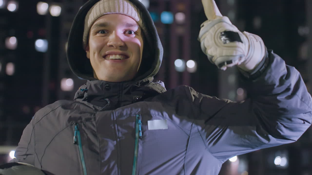 hombre emocionado con capucha sosteniendo una pelota de fútbol cerca de la cámara mientras sonríe brillantemente y da pulgares hacia arriba con la mano izquierda levantada por encima del hombro por la noche en un entorno urbano