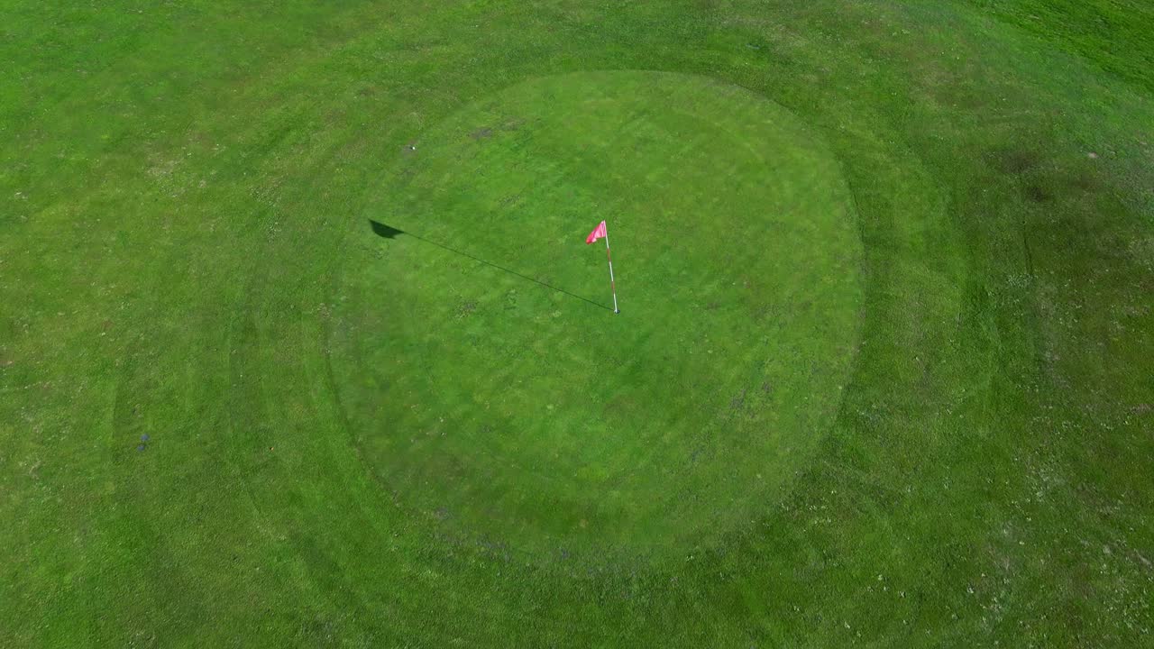 Backward vertical tilted up drone fly at grassy green environment with a single golf flag at the center