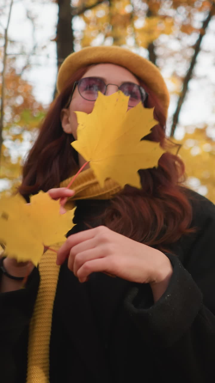 dama elegante con boina amarilla y silenciador juega sosteniendo la hoja de otoño cerca de la cara mientras pasea por un parque pintoresco, rodeado de follaje dorado, disfrutando de la naturaleza pacífica en el aire fresco de otoño