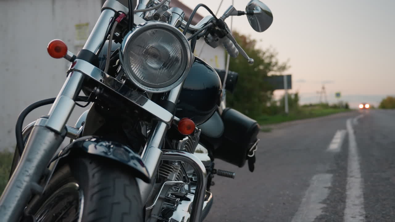 Parked motorcycle at roadside near small shelter with chrome handlebar details reflecting evening light while blurred car approaches on distant lane, creating cinematic sense of travel