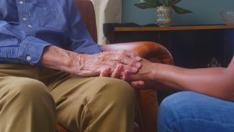 Close-Up-Of-Female-Care-Worker-In-Uniform-Holding-Hands-Of-Senior-Man-Sitting-In-Care-Home-Lounge