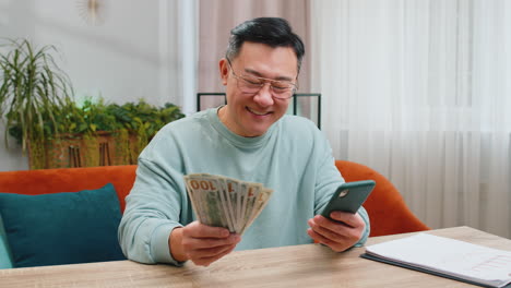 Rich-happy-Asian-man-counting-money-cash-on-smartphone-calculator-app,-calculate-income-earnings