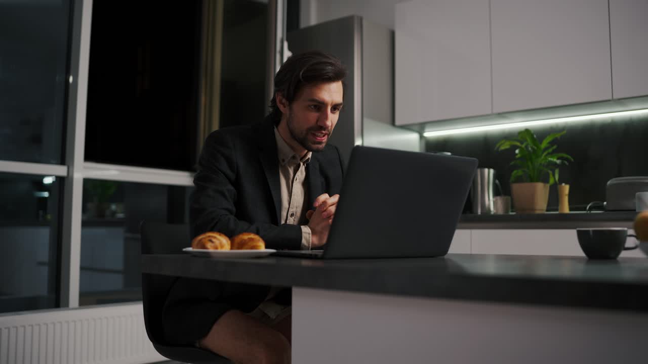 A serious and confident brunette man with stubble in a black jacket and beige shorts goes on a video call using a laptop for a video conference in the evening working remotely in a modern apartment