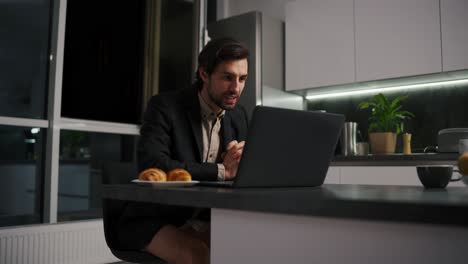 A-serious-and-confident-brunette-man-with-stubble-in-a-black-jacket-and-beige-shorts-goes-on-a-video-call-using-a-laptop-for-a-video-conference-in-the-evening-working-remotely-in-a-modern-apartment