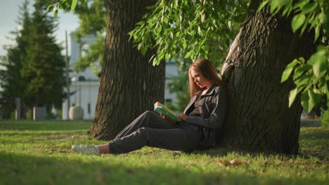 woman sitting outdoors with legs stretched, leaning against tree on grassy field, reading book, tree leaves sway gently in breeze, background featuring greenery and a white building