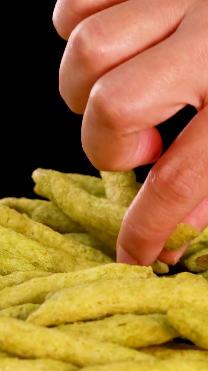 A hand selects a crispy green pea puffed snack from a pile against black background.