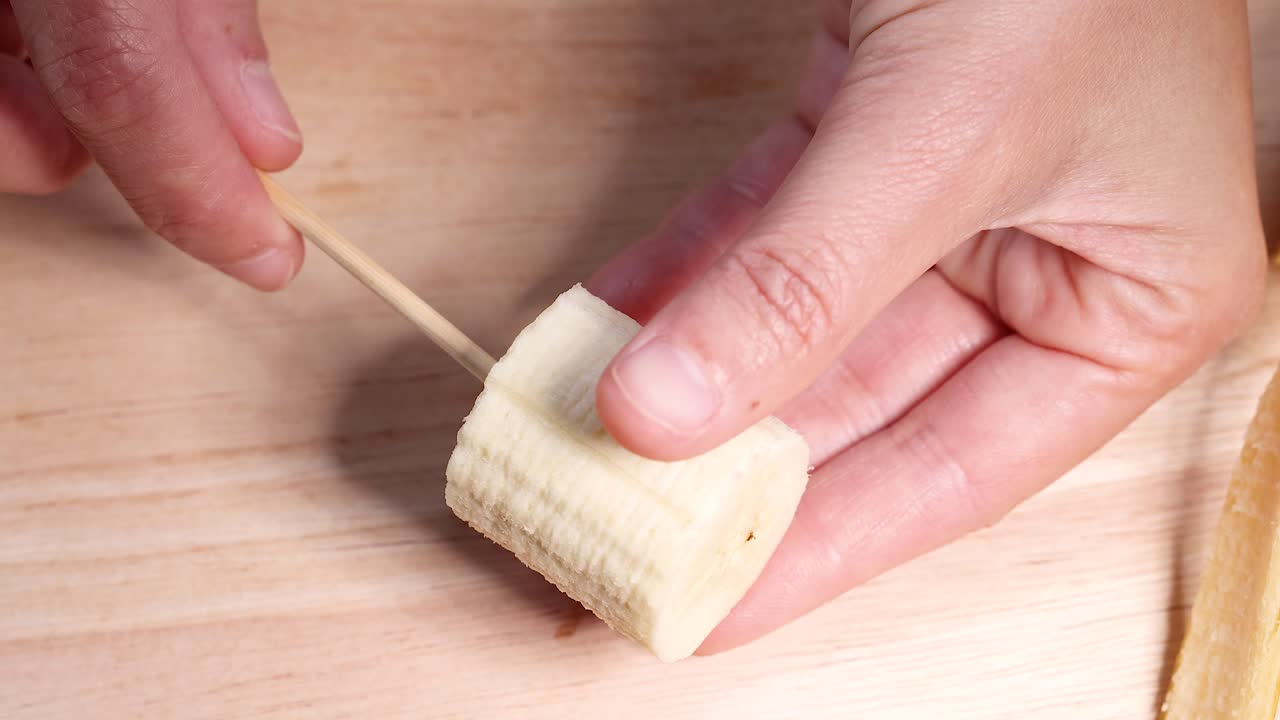 Hand skewers banana slice on wooden surface