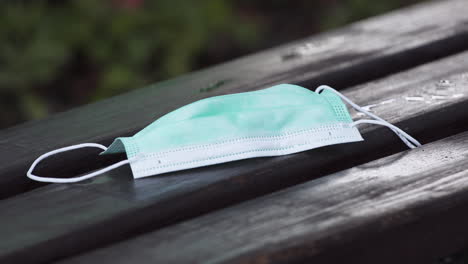 a person putting a simple green protective covid-19 face mask on a dark wooden bench, picking it up again after a moment, blurry bushes in the background, static close up 4k shot