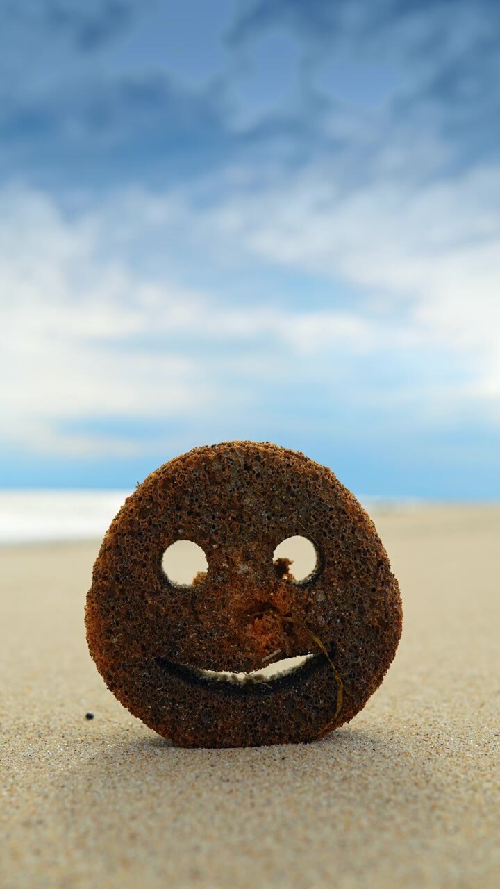 Smiley face cleaning sponge standing on a sandy beach. Circular cleaning sponge with a smiley face cut-out sitting on the sand with blurred ocean background.