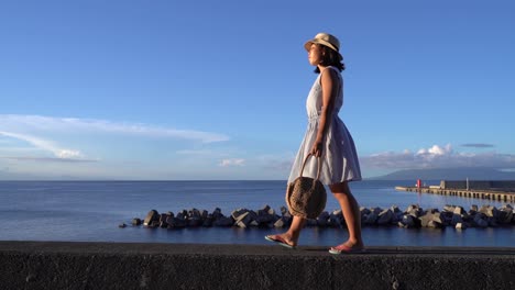 japanese girl in summer dress holding bag walking in front of ocean on beautiful clear day - wide sideways tracking shot slow motion