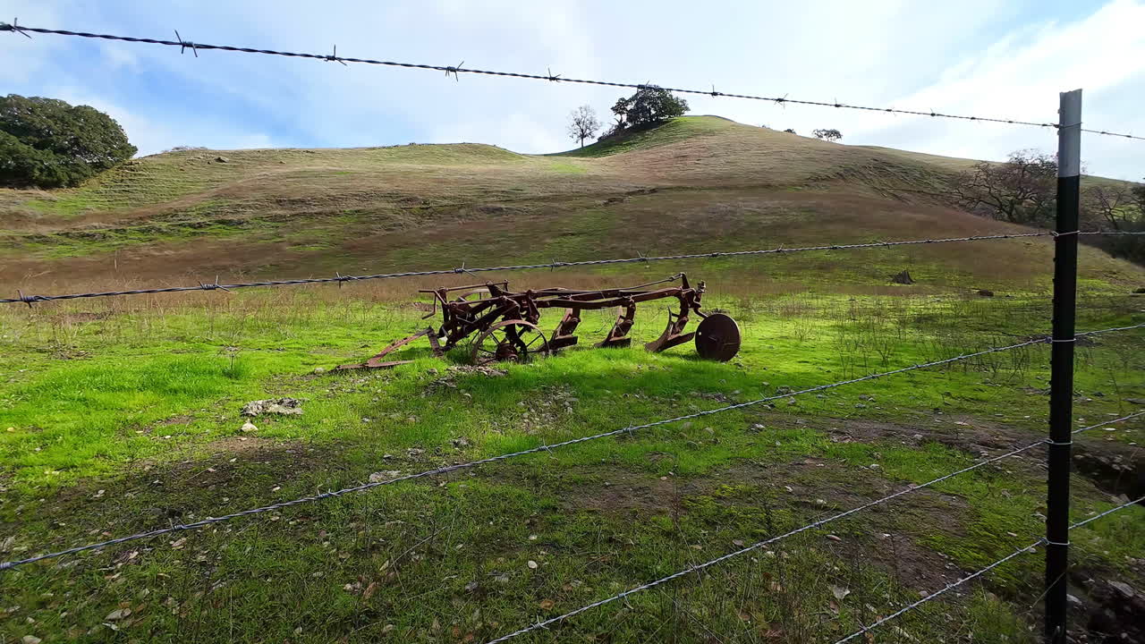 Drone flies through a barbed wire fence towards a meadow with an old plough