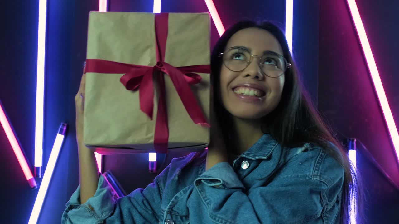 chica feliz con una caja de regalos y mirando a la cámara sobre el fondo de luz de neón