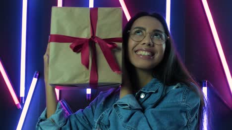 chica feliz con una caja de regalos y mirando a la cámara sobre el fondo de luz de neón