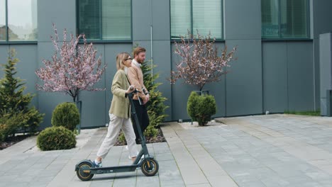 caucasian young woman and man walking on street with kick e-scooter and laptop talking and laughing by modern office buildings. technology, partnership, outside