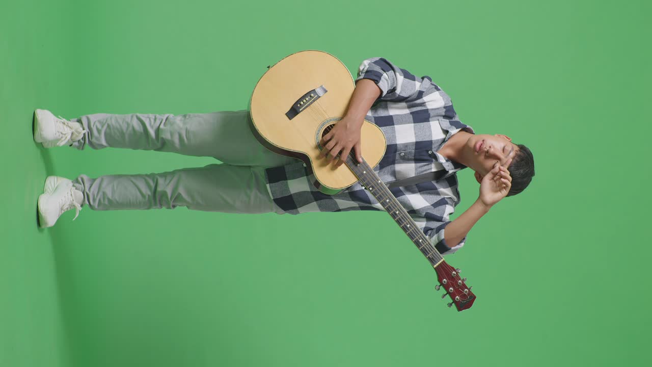 Figura intera di adolescente asiatico stanco con la chitarra in piedi su sfondo verde