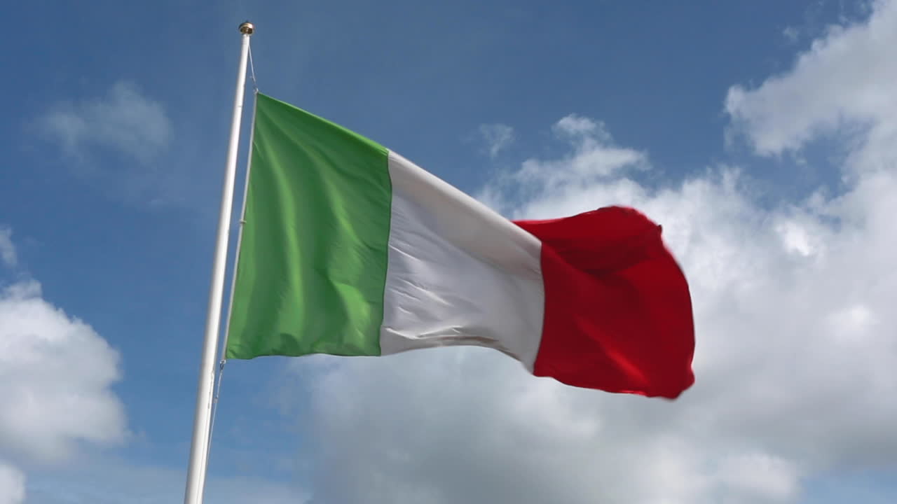 Bandiera italiana verde, bianca e rossa che sventola quasi dritta in un vento forte con un cielo blu reale e nuvole sullo sfondo