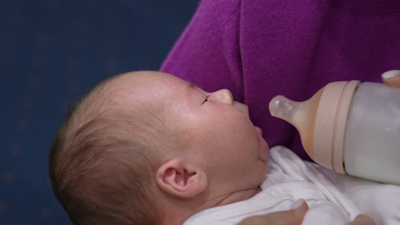 Baby lying in mommy's hands ready to be fed. Baby grasping the pacifier of the bottle and suckling hungrily. Close up.