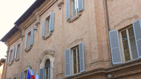 Low-angle-shot-of-historic-building-with-EU-and-Italian-flag-hanging-in-the-front-in-Urbino,-Italy-Old-town-architecture