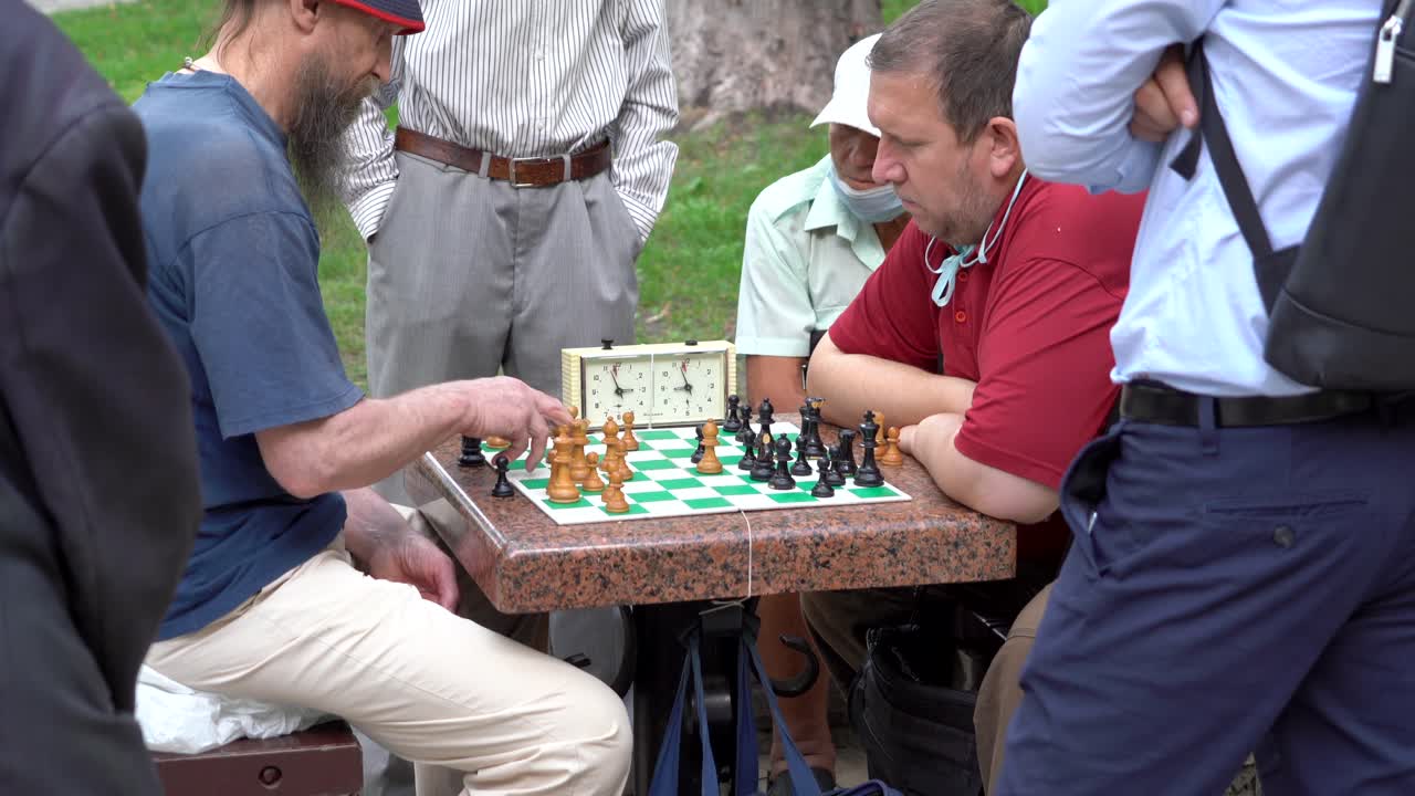 Ukraine, Kyiv, Two people playing chess at a park with a clock,others watching them while some others just walking on the park and enjoying the summer day.