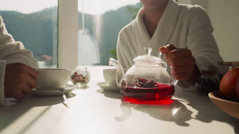 el hombre pone la tetera con el té en la mesa. los jóvenes disfrutan de la hora del té de la mañana sentados juntos en la cocina junto a la ventana. proceso de la ceremonia del té por la mañana