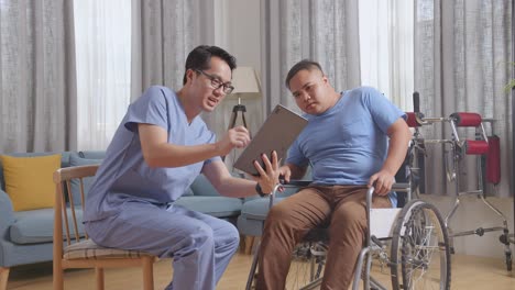 side view of asian male nurse showing medical test results on a tablet and explaining to a patient in wheelchair during a physical therapy at home