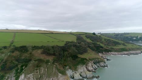 Aerial-ascending-forward-over-a-rocky-Devonshire-coastline-revealing-a-mixture-of-colourful-fields-inland