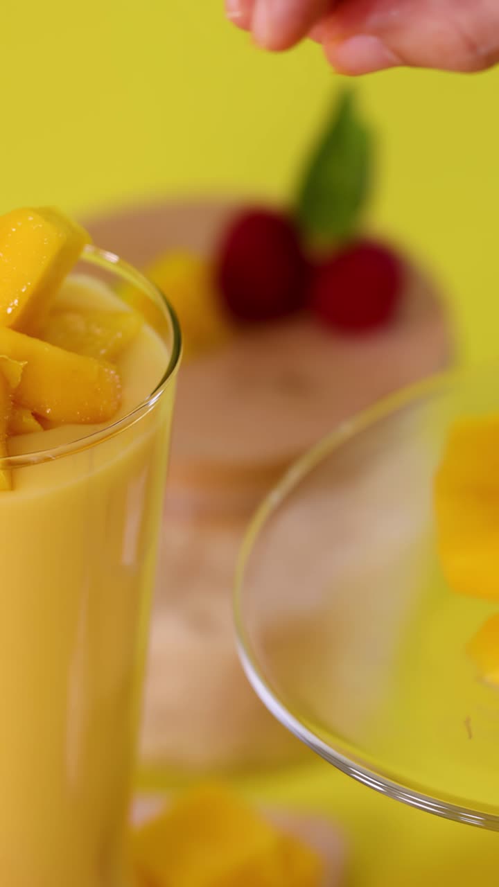 Primo piano di una mano che aggiunge cubetti di mango a un frullato in bicchiere su sfondo giallo brillante