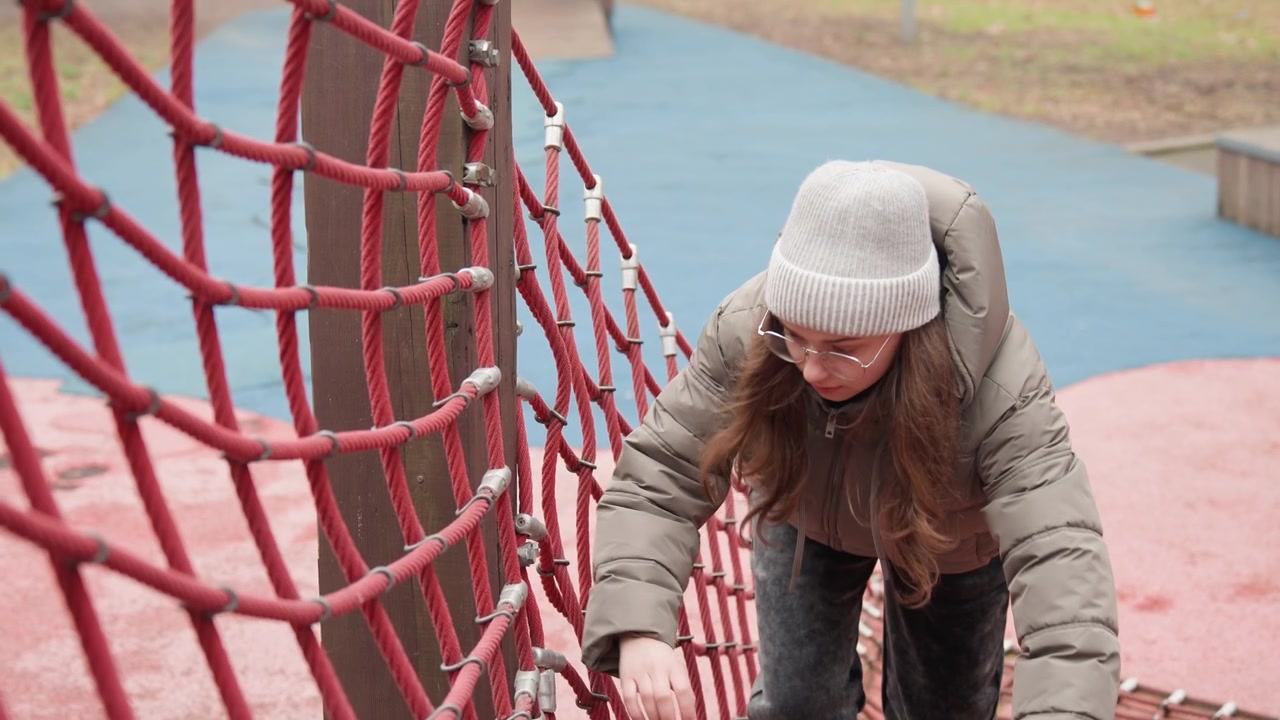 White woman balancing on rope net walking across suspended net playground winter coat jeans boots cautious strides hands gripping ropes dynamic movement focus determination outdoor training challenge
