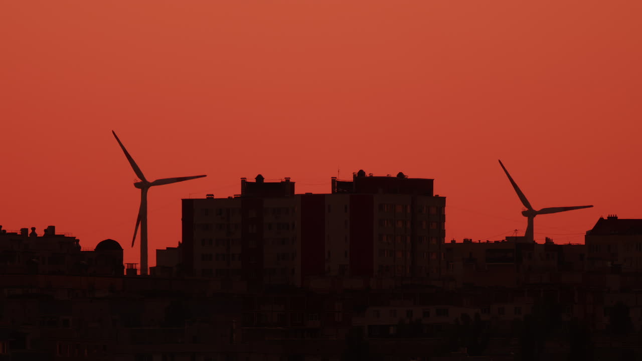 Two wind turbines stand tall near city buildings under an orange sky