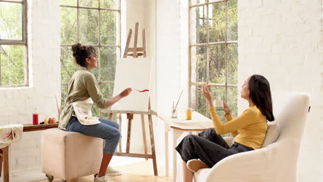 woman painting on canvas while friend enjoys coffee in bright home studio