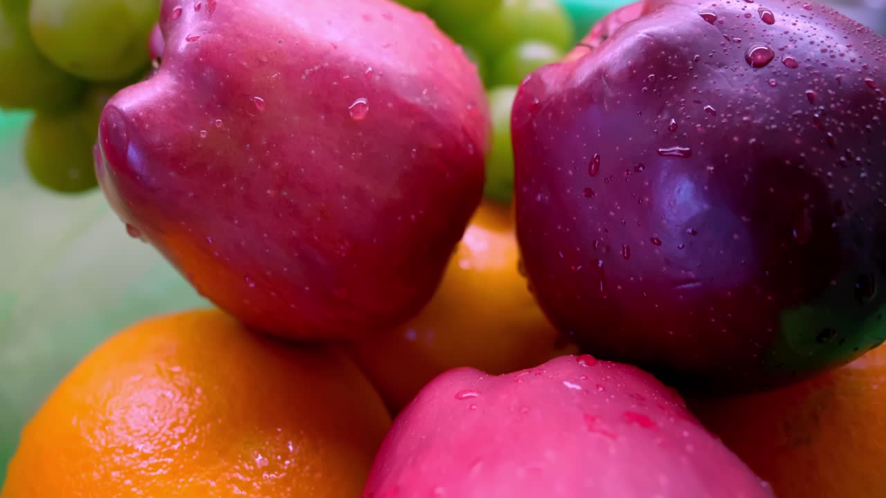 A bowl of fresh fruit with water droplets sits on a green surface, showcasing oranges, apples, and grapes