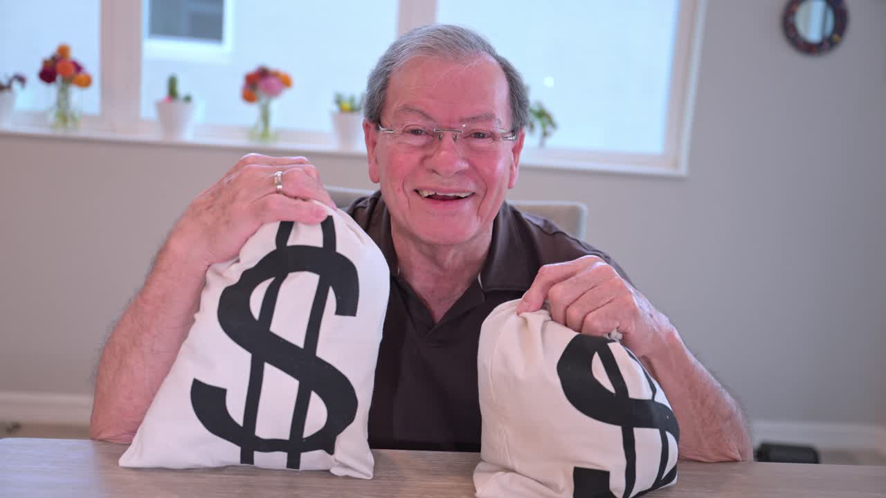 부유 한 노인 이 테이블 에 앉아 두 개 의 돈 가방 을 들고 미소 짓고 웃고 있다