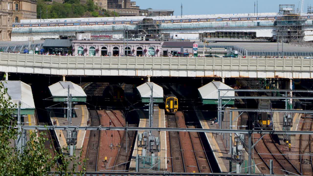 Yellow-fronted train approaches busy urban railway station under bright daylight, wide static shot