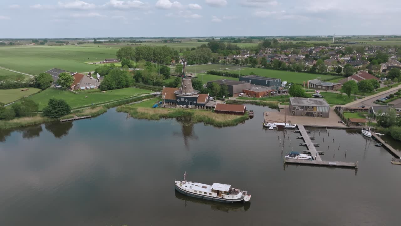 Drone zooms in toward a calm harbor with moored boats and village buildings surrounded by green fields in the Netherlands. In the background, a traditional windmill completes the idyllic landscape