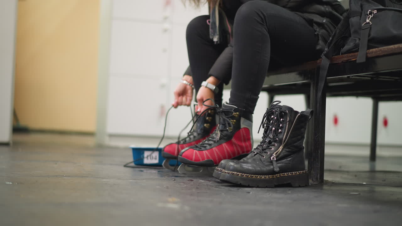 Lady preparing for skating by putting on red black ice skates while sitting on bench, wearing black pants and dotted socks, combat boots placed on floor beside, ready for winter sport in locker room