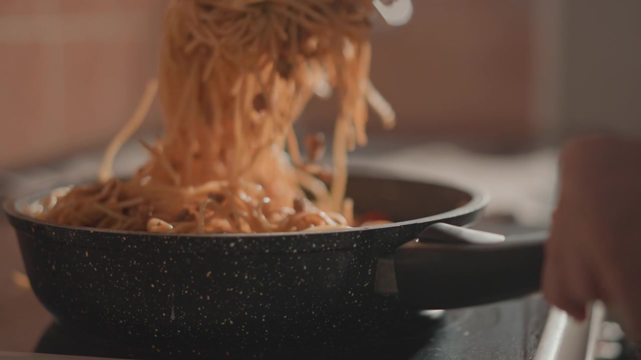 Le pinze sollevano e separano spaghetti lucidi da una padella nera maculata mentre un liquido cola sui noodles, con movimento verso l'alto, goccioline sospese e vapore leggero che aggiungono un tocco di freschezza e calore alla cucina.