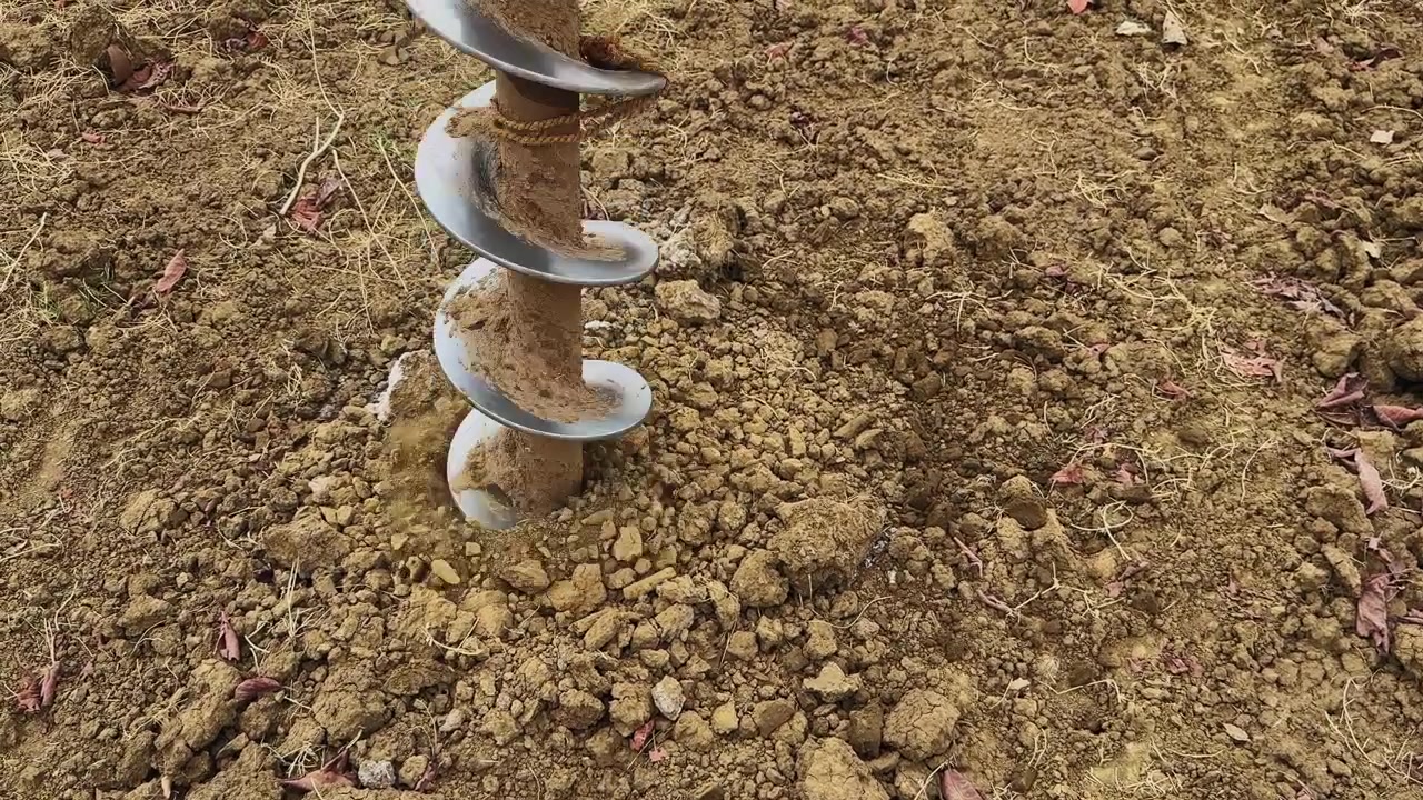 Mid view of auger drill bit partially inside ground with loose soil and gravel around, showing ongoing drilling process in a rural agricultural environment