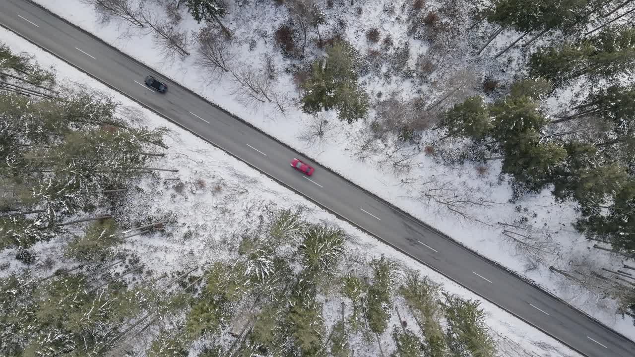 toma de un dron de una carretera enmarcada por la nieve que conduce diagonalmente a través de un bosque con dos autos conduciendo, una toma de invierno de arriba hacia abajo