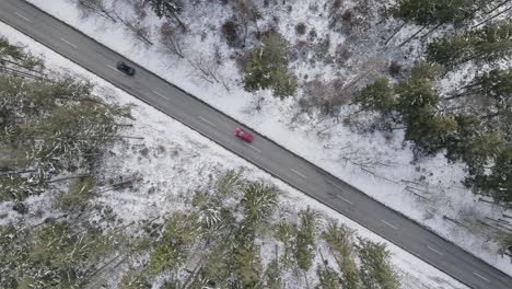 Drone-shot-of-a-diagonal-leading-snow-framed-road-through-a-forest-with-two-driving-cars,-a-top-down-winter-shot